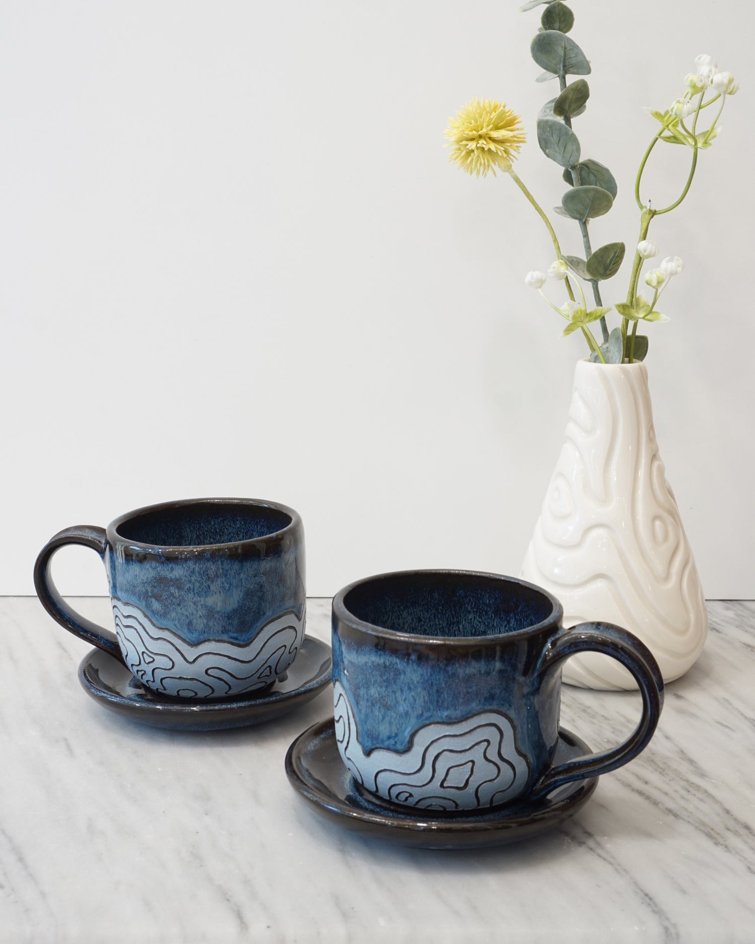 Image showing two espresso cups with saucers with a topographic map design with a white porcelain vase and flowers also with a topographic map design on a marble counter with white background
